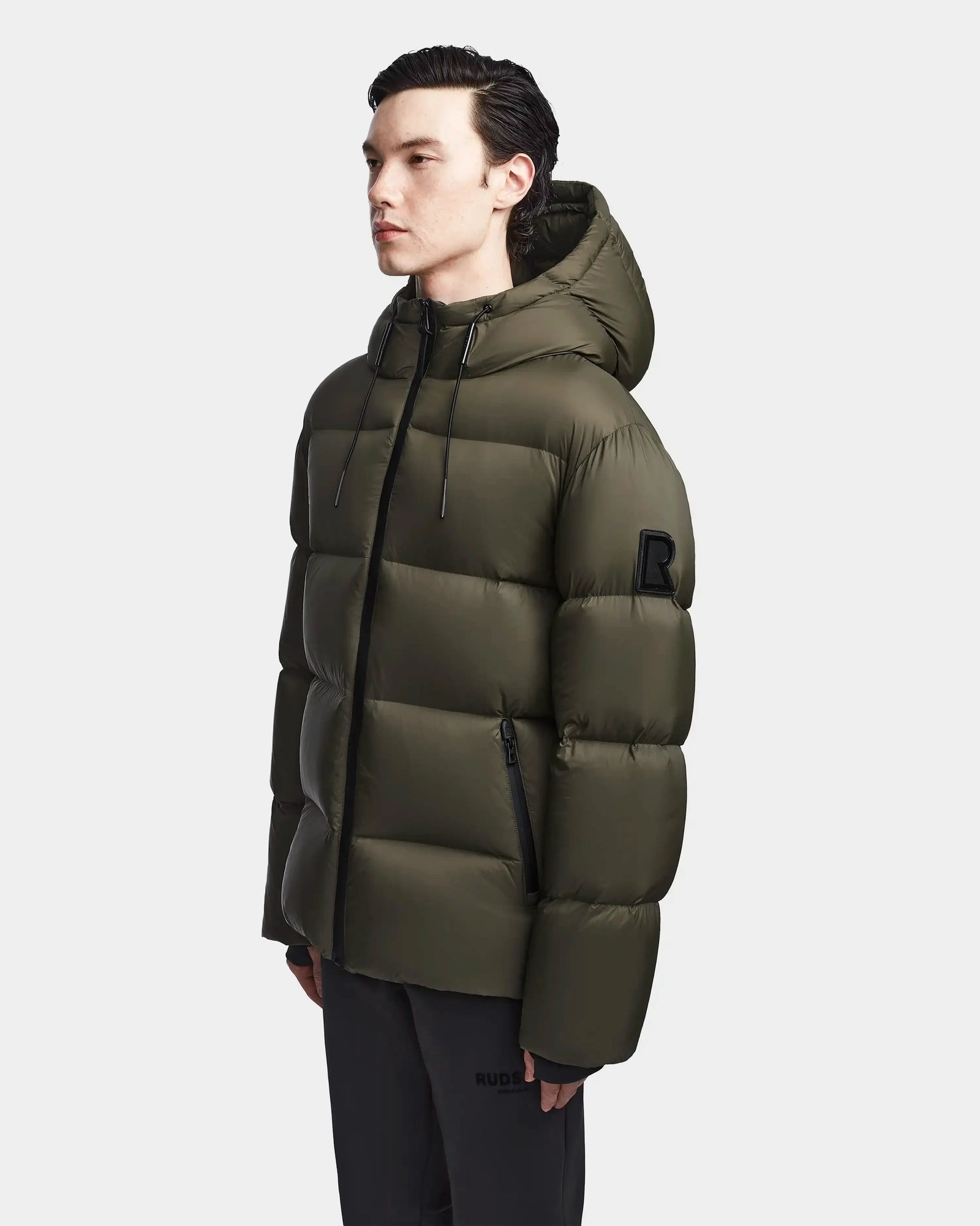 A man wearing the Rudsak DANIEL M - M123510 Light Olive mens hooded down puffer with a black zipper and upper arm patch stands facing left against a plain light background.