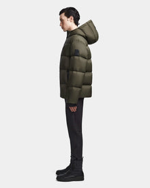 A person stands in profile against a plain background, wearing the Rudsak DANIEL M - M123510 Light Olive mens coat with a hood, paired with black pants and black lace-up boots.