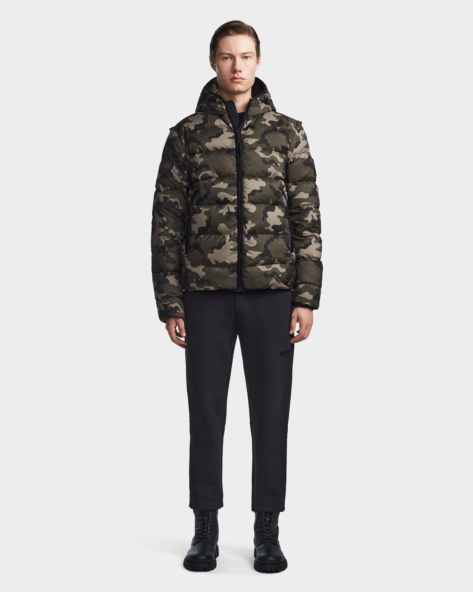 A person stands facing forward wearing the Rudsak DEMIAN M-R - M123504 Camouflage mens down puffer, paired with black pants and black lace-up boots against a plain light background.