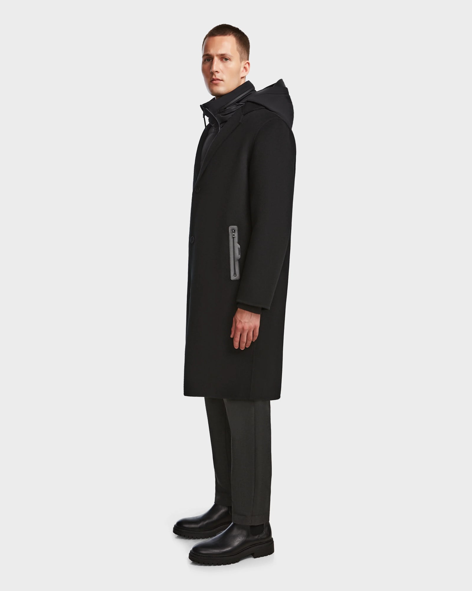 A man stands sideways against a plain white background, wearing the Rudsak DRAL - 6124503 Black mens wool jacket with a hood, paired with dark trousers and black boots.
