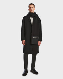 A man stands against a plain background wearing the Rudsak DRAL - 6124503 Black wool jacket styled as a long coat, paired with a black scarf, gray trousers, black boots, and holding a black zippered pouch.