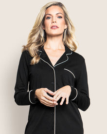 A person models the Womens Pima Nightshirt in Black by Petite Plume, featuring white piping and a chest pocket, reminiscent of luxury pajamas. They have long, wavy blonde hair and are posed against a light backdrop, gazing into the camera.