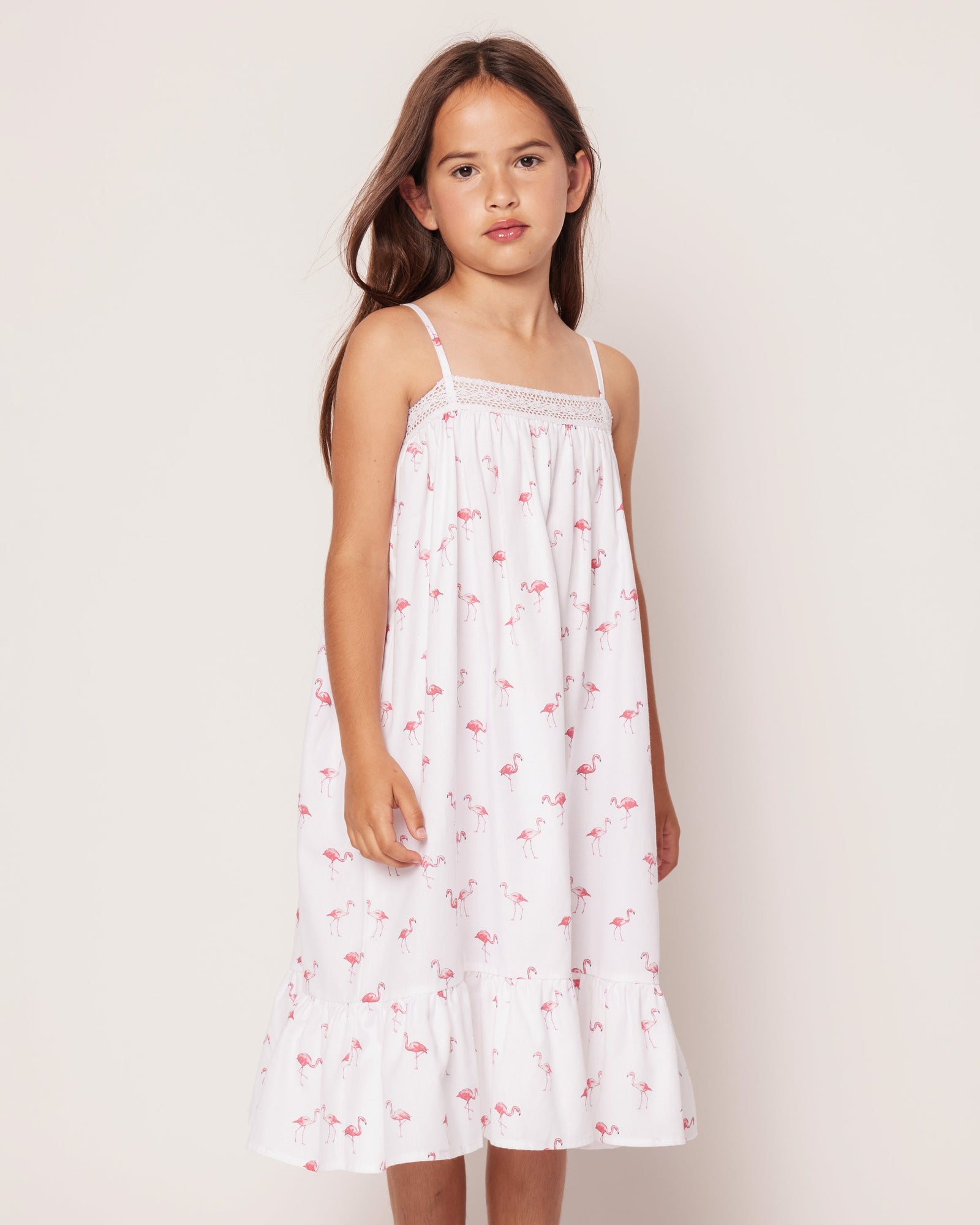 A young girl with long brown hair wears Petite Plumes Girls Twill Lily Nightgown in Flamingos, featuring thin straps and a ruffled hem. She stands against a plain off-white background with a neutral expression, the small pink flamingo pattern adding a tropical twist.