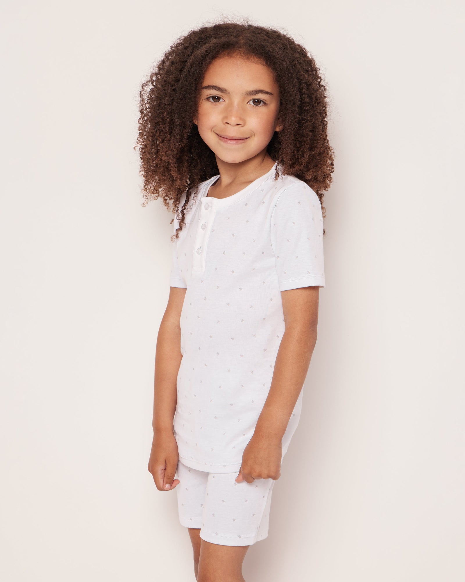 A young child with curly hair smiles against a light background, dressed in Petite Plumes Kids Pima Snug Fit Pajama Short Set in Grey Stars. The white short-sleeved shirt and shorts feature a subtle pattern and neckline buttons, crafted from soft Pima Cotton for comfort.