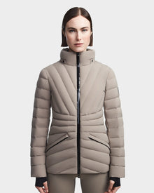 A woman with straight brown hair wears the Rudsak HANNA - 8123562 Cement, a light beige down jacket with a high collar and black zipper, standing forward against a plain white background.