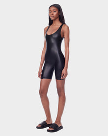 A woman wears the Rudsak AVERI - 8523015 BLACK fitted, shiny, sleeveless romper with shorts and black slide sandals against a plain white background.