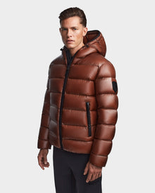A man wearing the Rudsak DEMIAN S - M123508 Cohiba, a shiny brown men’s down puffer with hood and black zippers, stands against a plain background. This water- and wind-resistant jacket blends style with functionality.