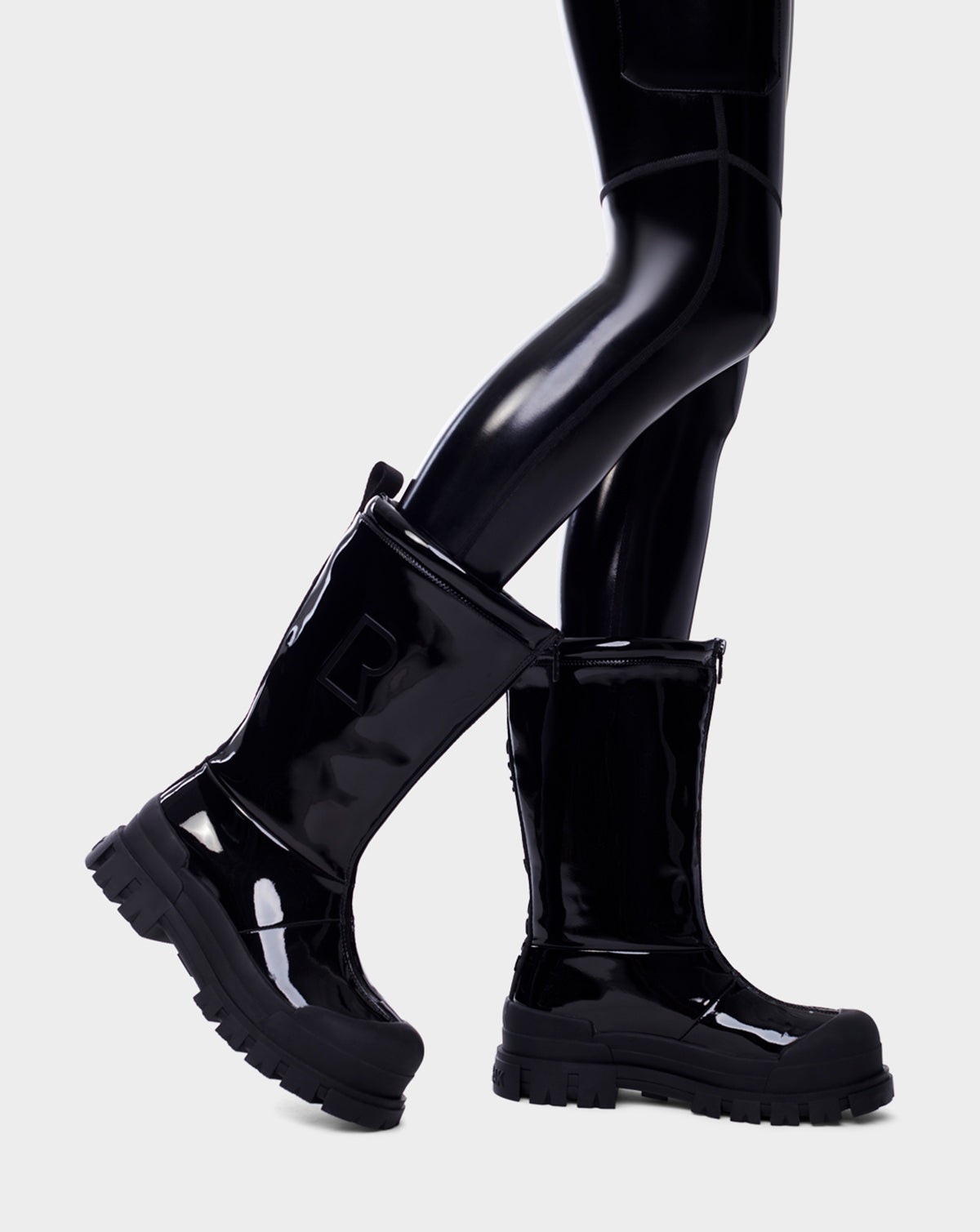 A person wearing Rudsaks KRISTAL HI - 8222510 BLACK high-gloss womens boots with chunky soles and black leggings stands with one foot slightly raised against a light gray background.