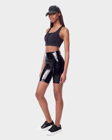 A woman in Rudsak’s BRIA - 8523016 BLACK vegan leather biker shorts, black crop top, sneakers with white soles, and a black cap stands against a light gray background, showcasing sustainable fashion.