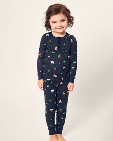 A young child smiles, clad in Petite Plumes cozy Kids Pima Snug Fit Pajama Set in Arctic Antics. The long sleeve navy blue top, adorned with small animal and forest prints, pops against a plain light background.