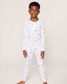 A young boy smiles, wearing the Petite Plume Kids Pima Snug Fit Pajama Set in Jingle Paws. This luxurious sleepwear features colorful small animal prints and provides comfort with its long sleeve top and pants as he stands barefoot against a plain background.