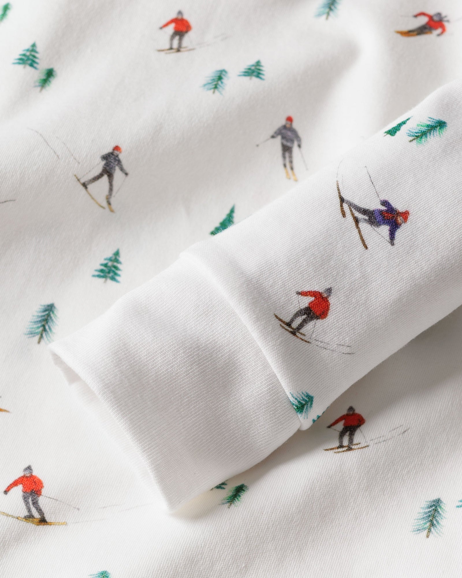 A close-up of Petite Plumes Apres Ski Kids Pima Snug Fit Pajama Set showcases skiers in vibrant attire, moving in various directions on soft and cozy white fabric, dotted with small green pine trees—ideal for a snug long sleeve top or pant set.
