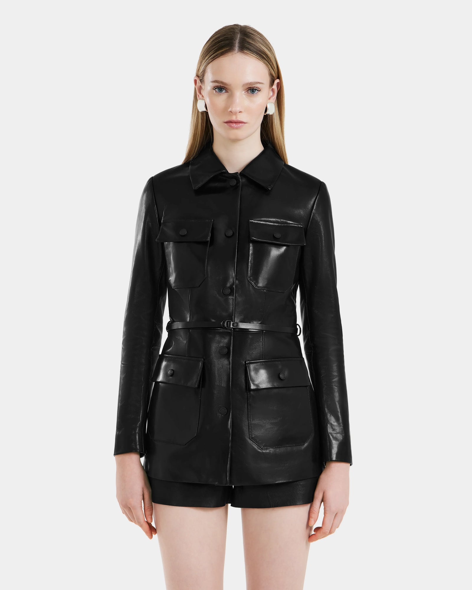 A woman with straight, long blonde hair models the Rudsak DANICA - 8125025 Black leather jacket, featuring four front pockets and buttons, paired with black shorts against a plain white background.