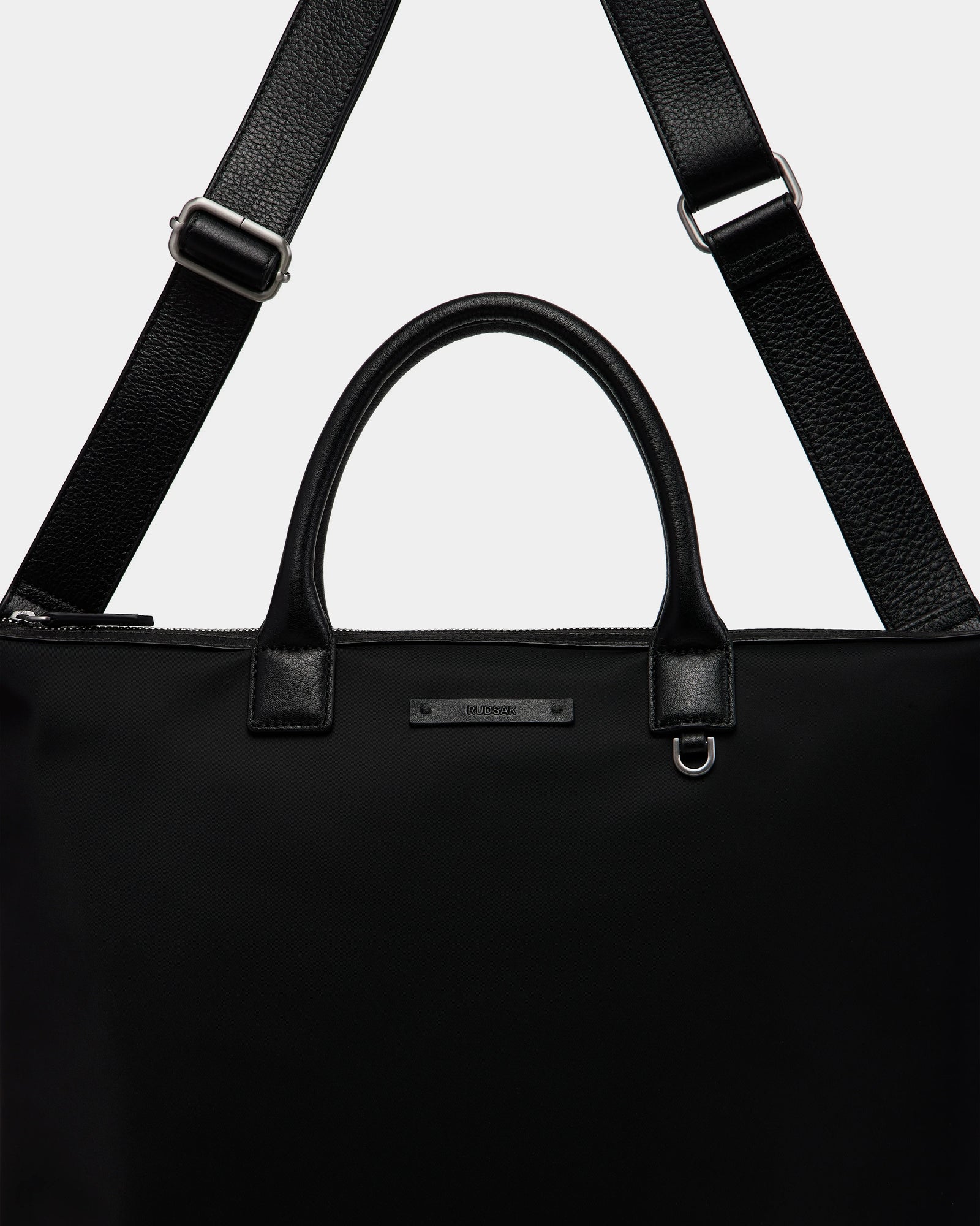 A close-up of the Rudsak DEXTER - 9324513 Black tote, a sleek unisex bag with sturdy leather handles, a detachable shoulder strap, and a small metallic brand label on the front.
