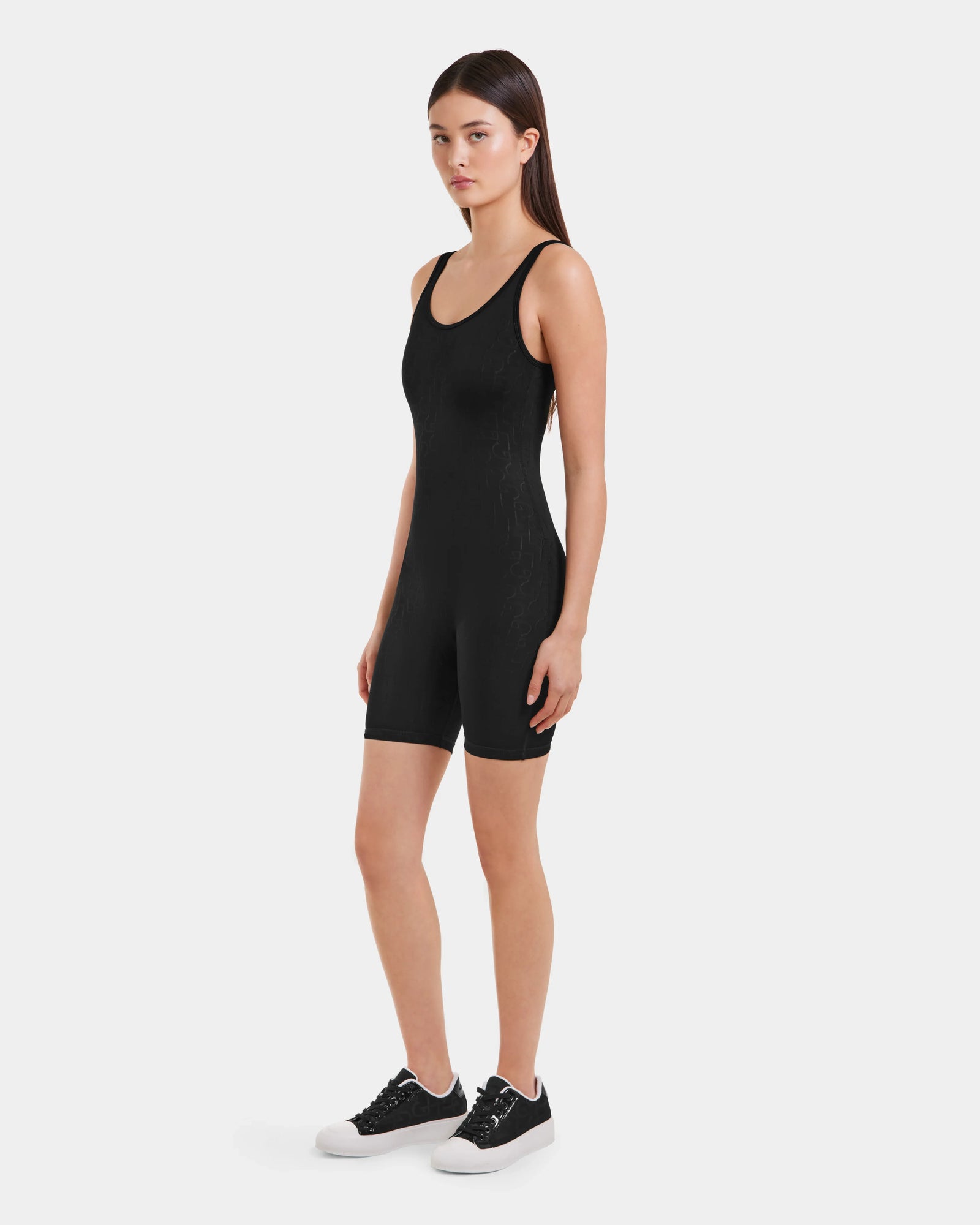 A woman with long, straight hair and a neutral expression stands against a white background wearing the Rudsak KAILEY - 8523012 Black Pattern fitted sleeveless bodysuit romper and black sneakers, facing slightly to the side.