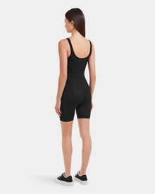 A woman with straight brown hair faces away, wearing the Rudsak KAILEY - 8523012 Black Pattern bodysuit and black sneakers with white soles, set against a plain white background.