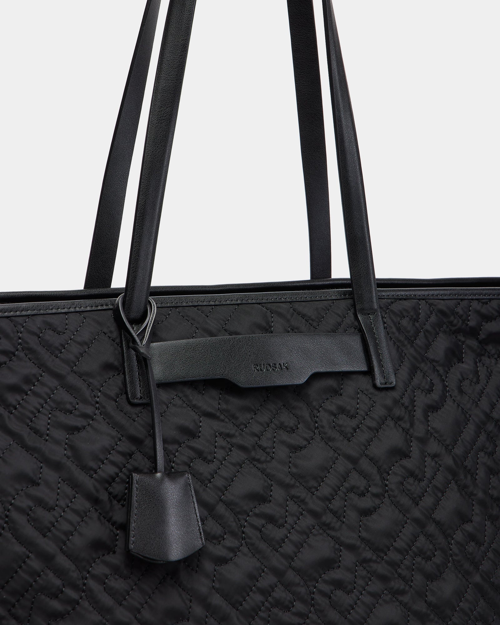 Close-up of the Rudsak LIVORNO - L325007 Black quilted monogram tote with embossed patterns, two long handles, a small leather tag, and a subtle brand name embossed on a leather strip near the top.