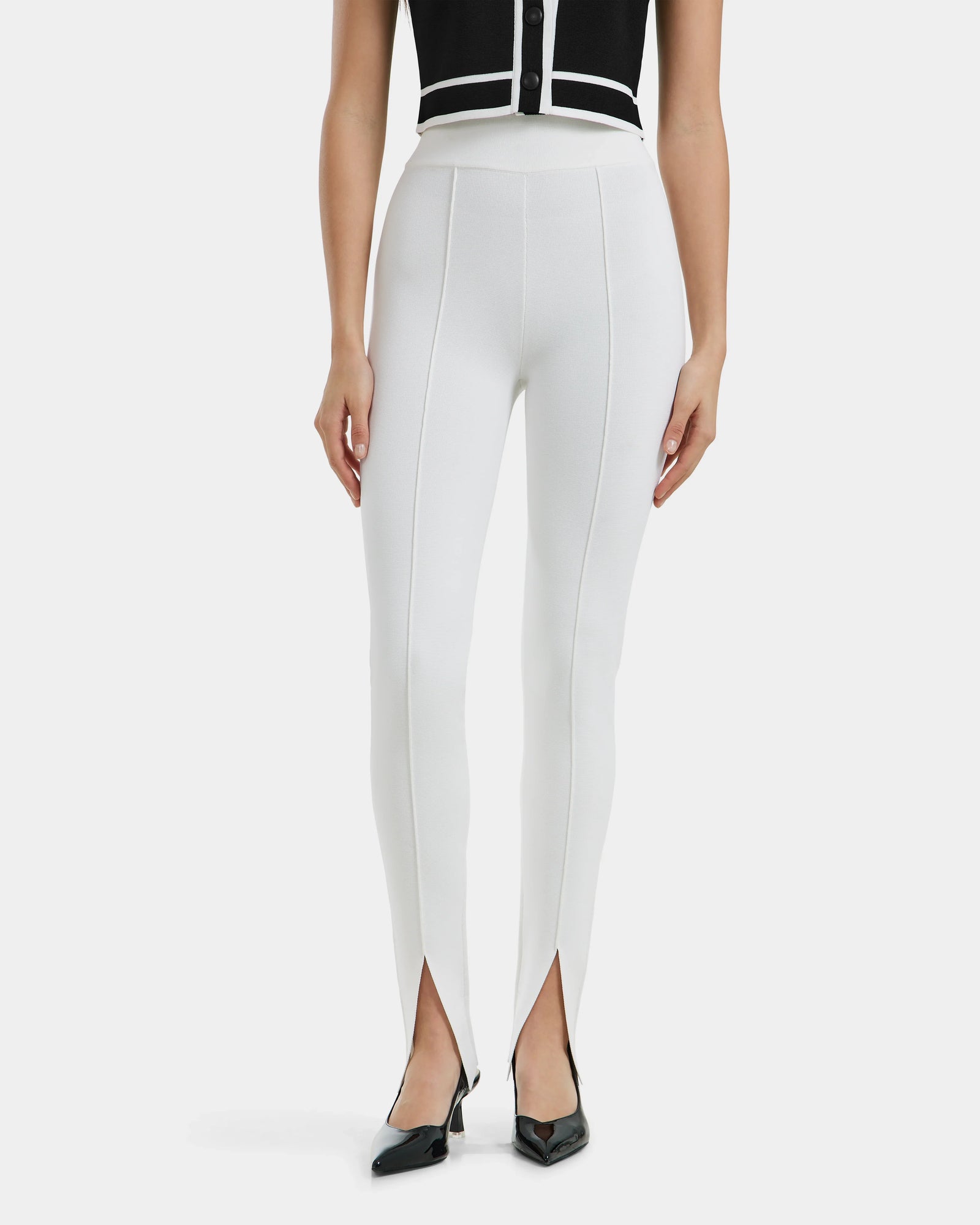 A woman wears Rudsak’s MATHILDA - 8524515 Iceland high-waisted white ankle-slit leggings with front center seams and split hems, styled with black pointed-toe heels and a sleeveless black top.