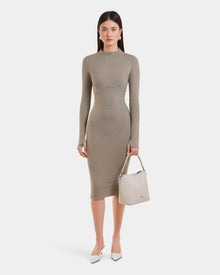 A woman stands against a white background wearing the Rudsak MAYLIN - 8523504 Cement Pattern women’s long fitted dress, paired with white pointed-toe heels and holding a beige handbag in her left hand.
