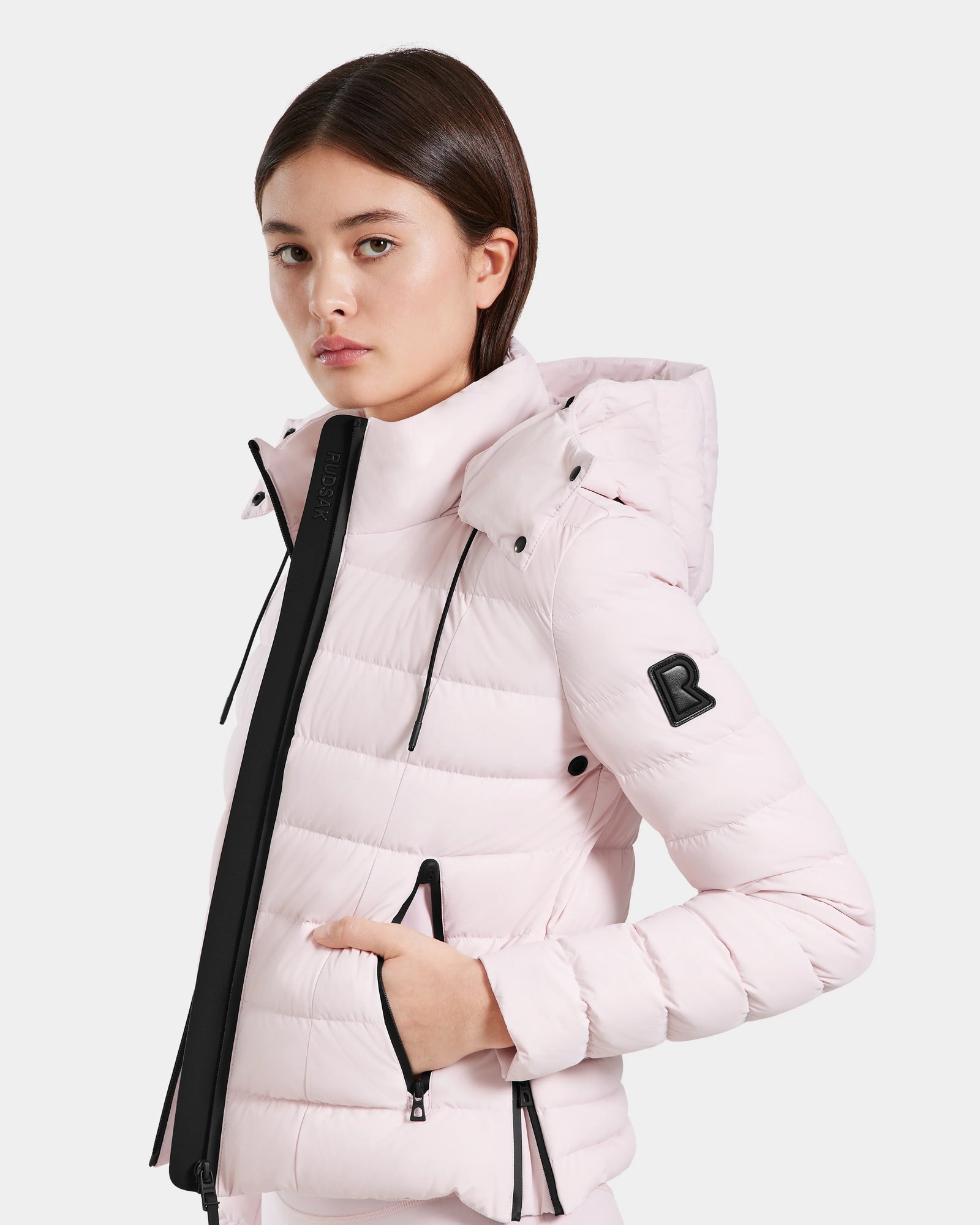 A young woman models the Rudsak MELISSA - 8125054 Light Pink puffer jacket, featuring a hood, black zipper accents, and a signature “R” patch on the sleeve—highlighting this stylish, water-resistant outerwear against a plain background.