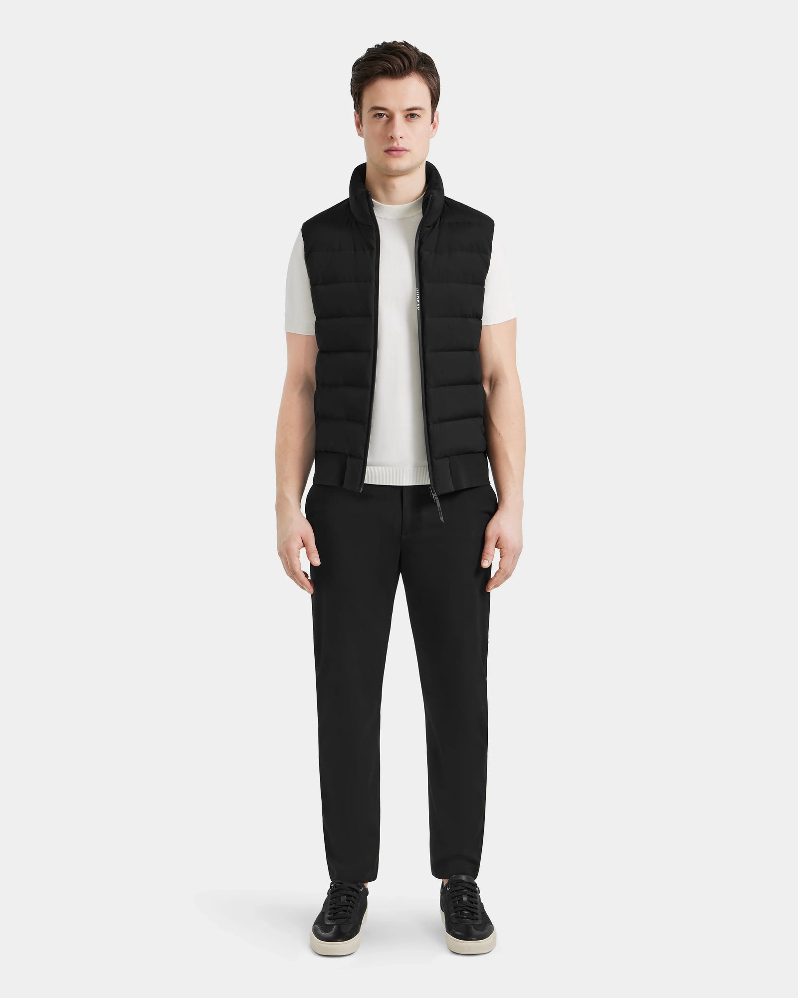 A man faces forward against a light gray background, wearing the Rudsak MERLO - 6124017 Black quilted vest over a white t-shirt, paired with black pants and black sneakers with white soles.