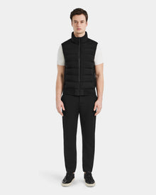 A man faces forward against a plain white background, wearing the Rudsak MERLO - 6124017 Black quilted vest over a white short-sleeve shirt, paired with black pants and black sneakers with white soles.
