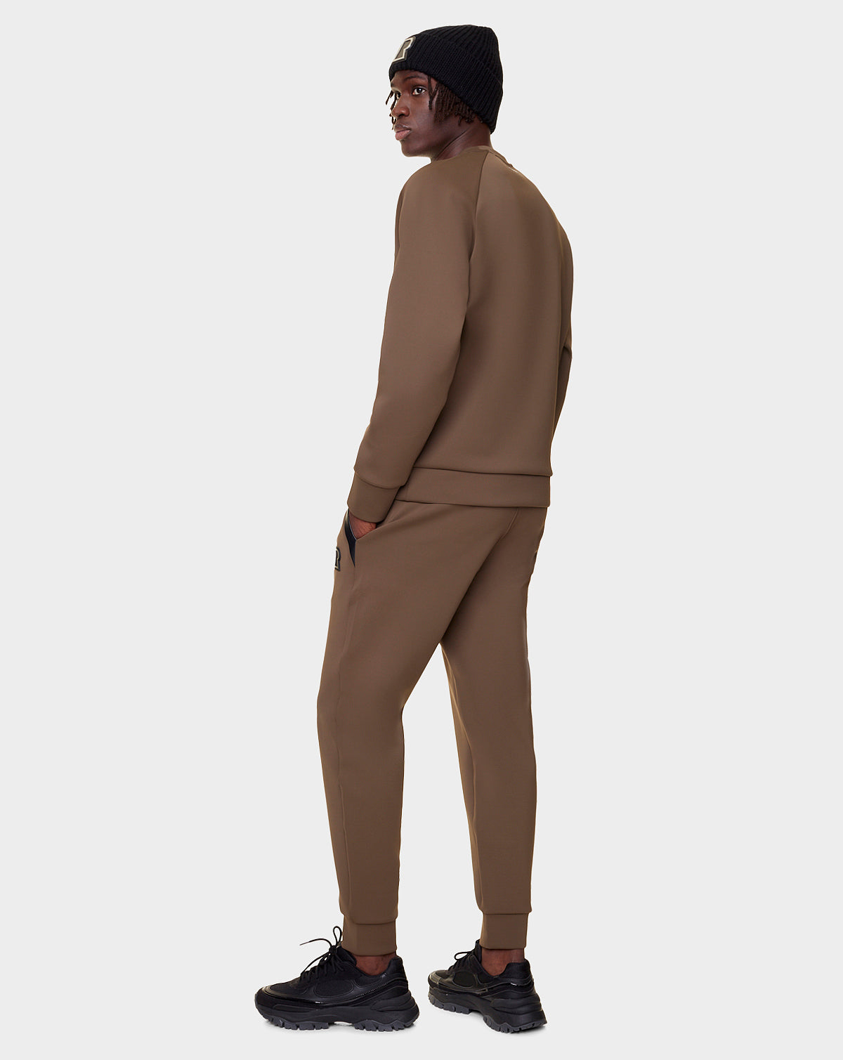 Wearing the Rudsak DEVAN - 9522510 ARMY MEN relaxed fit unisex crewneck sweater and matching jogger pants, paired with black sneakers and a black knit beanie, a person stands facing away, looking over their shoulder against a light gray background.