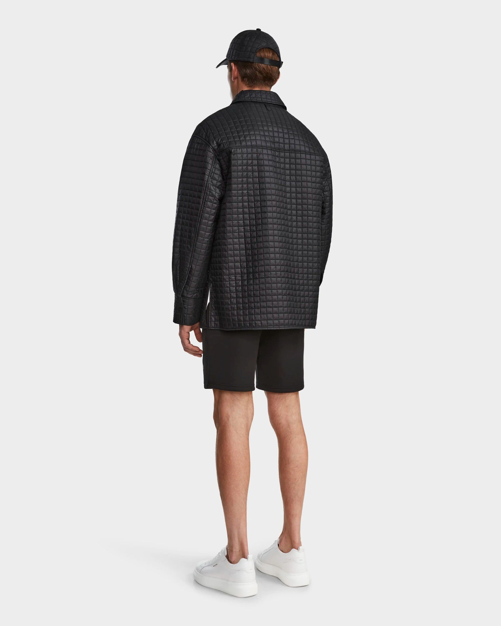 A man faces away in a Rudsak KERRY - 9124003 Black M quilted shirt jacket with eco-friendly insulation, paired with black shorts, black cap, and white sneakers against a plain white background.