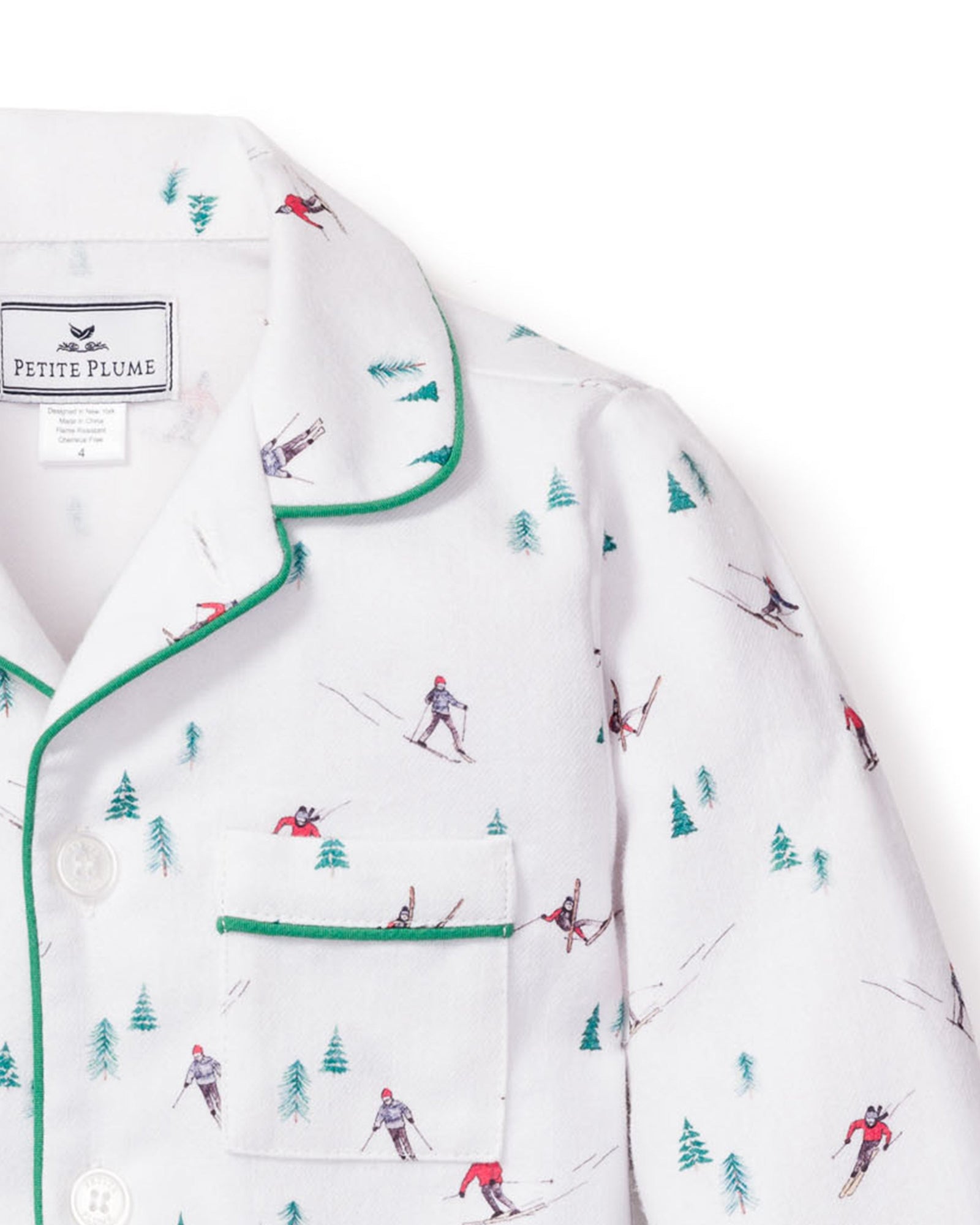 Petite Plumes Kids Flannel Pajama Set in Apres Ski features white flannel adorned with skiers and green trees, green piped trim, and a chest pocket. The collar label assures quality comfort with flame-retardant fabric.