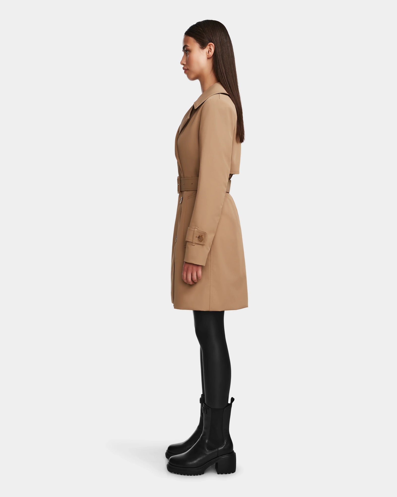 A woman in profile wears the Rudsak KIMBERLY - 8124801 Camel trench coat, black leggings, and chunky black boots against a plain white background.