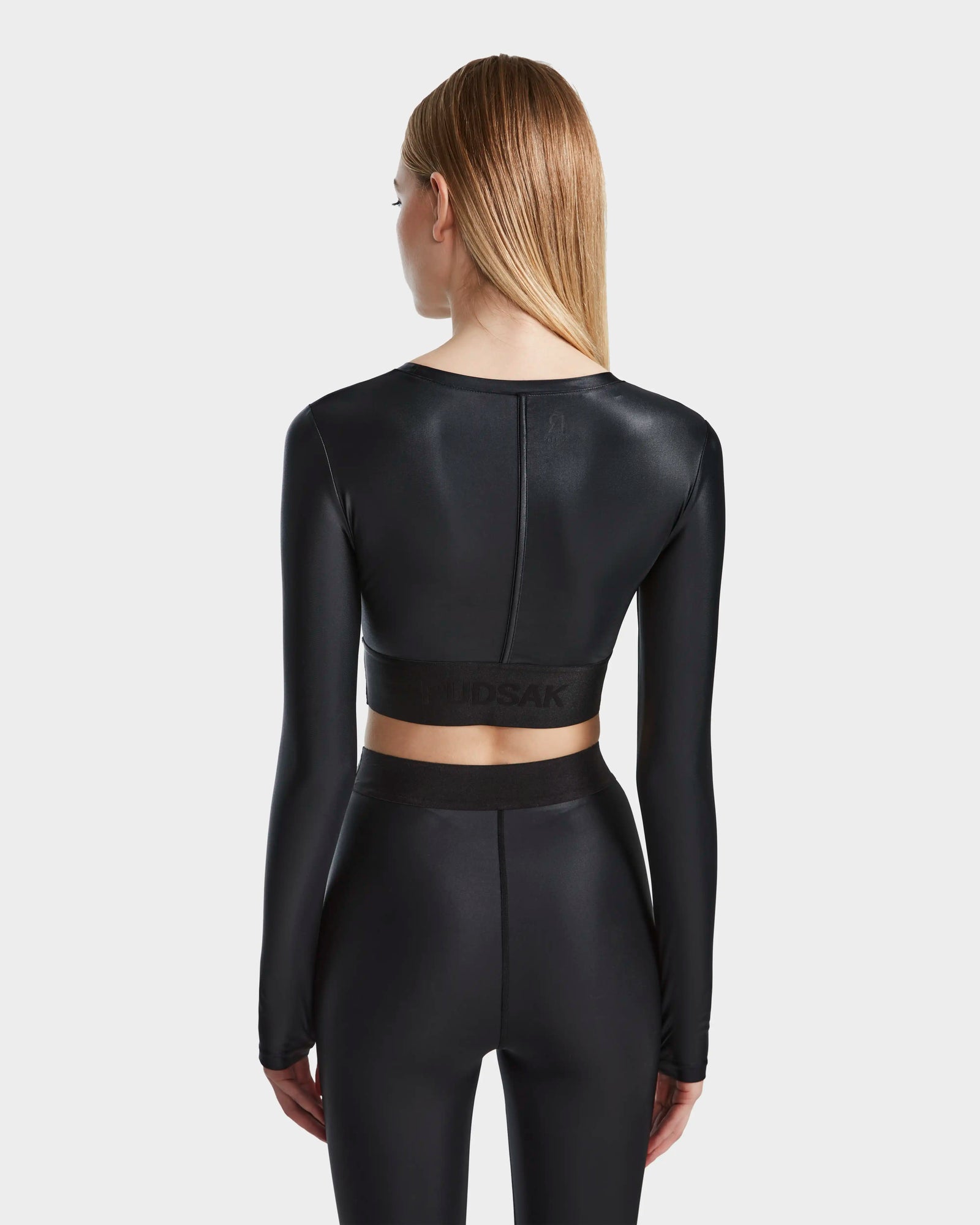 A woman with straight, light brown hair stands facing away, wearing the Rudsak LORA - 8524026 Black fitted long sleeve top and matching high-waisted leggings against a plain white background.