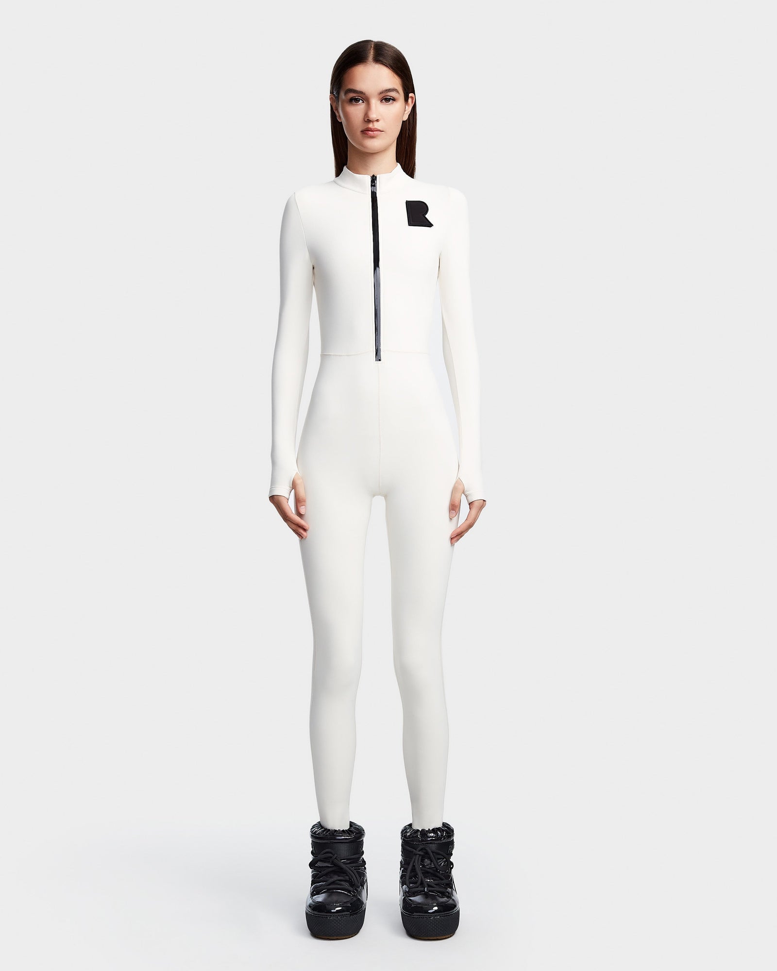 A woman models the Rudsak DAVINA - 8523534 Iceland: a sleek, white, long-sleeve jumpsuit with front zipper and black R on the chest, styled with chunky black boots against a plain white background for an après-ski look.
