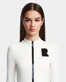 A young woman with straight brown hair models the Rudsak DAVINA - 8523534 Iceland: a fitted, fleece-lined bodysuit with long sleeves, black zipper, and bold black R patch, evoking après-ski style against a plain light grey background.