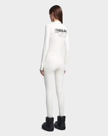 A woman with straight brown hair stands facing away in the Rudsak DAVINA - 8523534 Iceland fitted, fleece-lined jumpsuit. She wears black winter boots and channels ski-wear luxury against a light gray background.