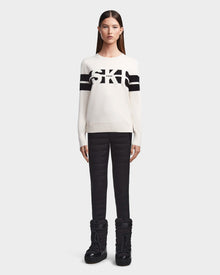 A woman faces forward in Rudsaks ODESSA - 8523540 Iceland soft-knit après-ski sweater with black stripes and “SKI” across the front, paired with black quilted pants and snow boots—ideal winter warmth.