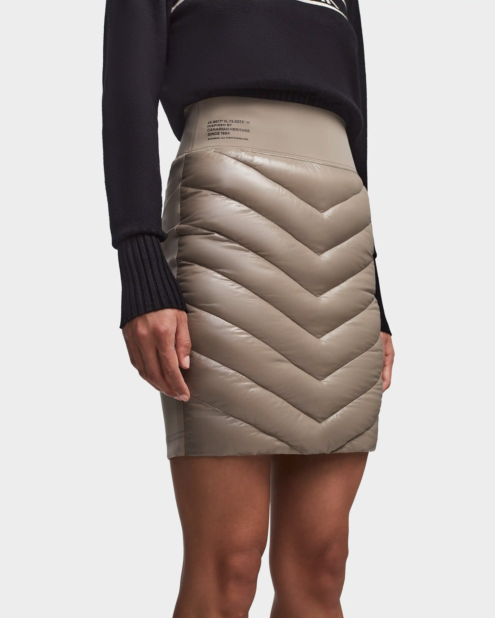 A person wearing the Rudsak MARZI - 8523528 Cement skirt, a shiny chevron-quilted taupe winter piece with a high waist, styled with a long-sleeve black sweater. The photo is cropped at the shoulder and mid-thigh.