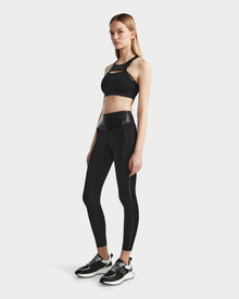 A woman poses in the ARYA - 8523002 BLACK sports bra and matching high-waisted leggings by RUDSAK, paired with black sneakers with white soles, set against a plain light background.
