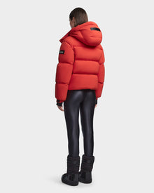 A woman in the Rudsak MAVE M - 8123572 Orange down puffer jacket with a hood, shiny black leggings, and black snow boots stands facing away from the camera against a plain white background.
