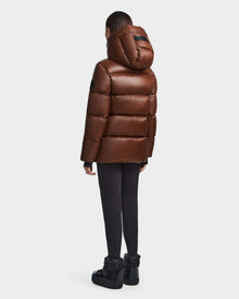 A person stands facing away, wearing the Rudsak RITA S - L123527 Cohiba shiny brown women’s hooded down puffer jacket, paired with black leggings and black platform shoes, against a plain white background.