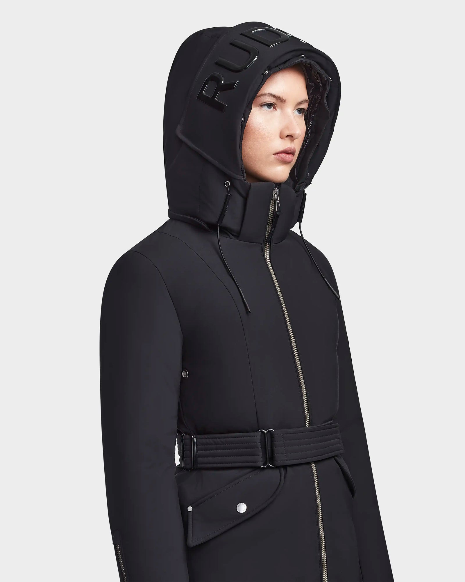 A person models the MILENA - 8122536 BLACK women’s winter coat by RUDSAK, featuring a belted waist, metallic zipper, and a long down parka hood with bold RUDSAK lettering for a chic look.