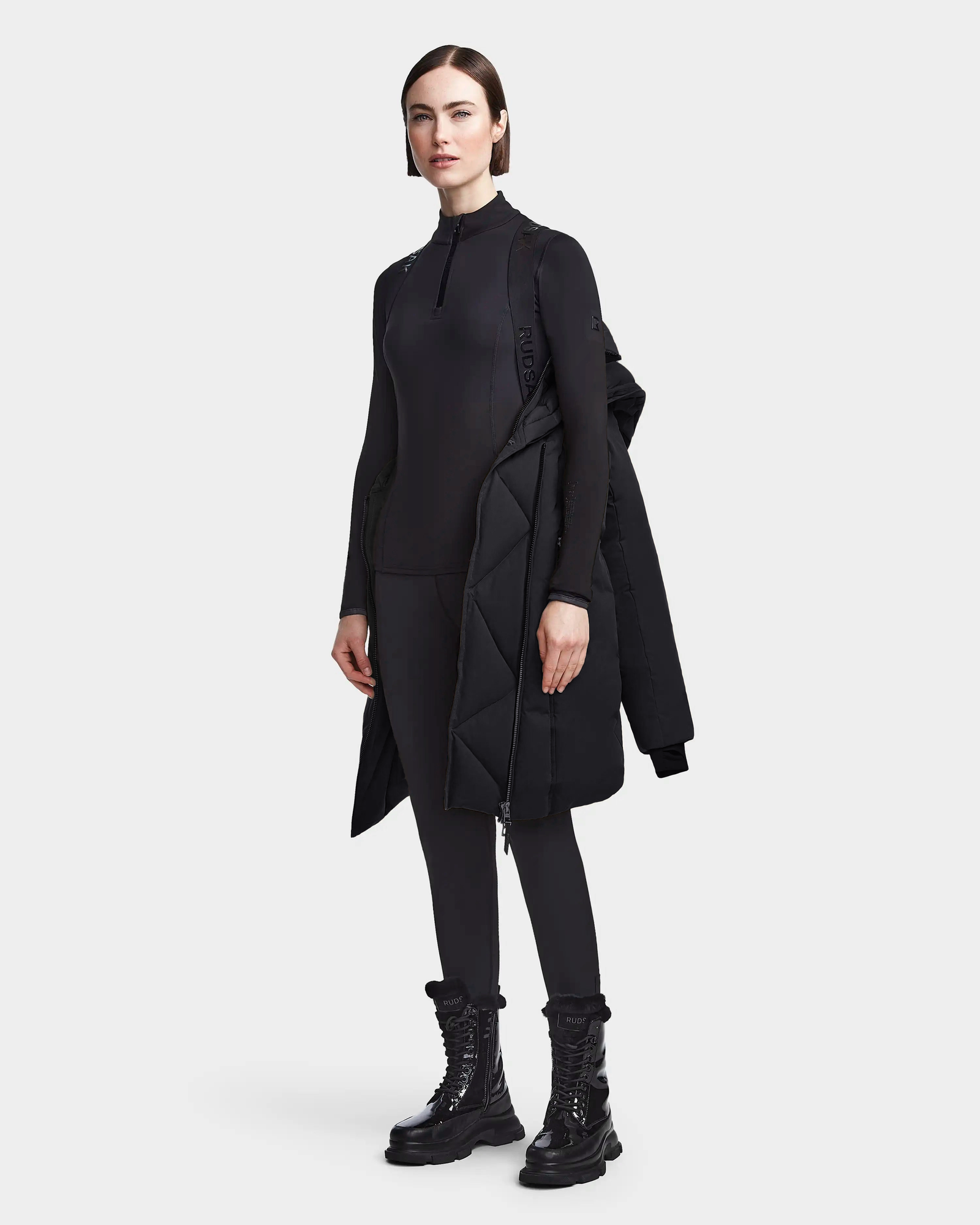 A woman with straight dark hair and a neutral expression stands against a plain background wearing Rudsaks MAIA M - 8123582 Black womens down parka with a faux fur hood, styled over an all-black outfit with lace-up boots.