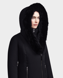 A woman wears the Rudsak MAIA M - 8123582 Black down parka with a large, fluffy faux fur hood, looking slightly to the side against a plain light background.