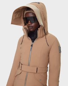 A person wears a RUDSAK MILENA - 8122536 TOFFEE womens toffee parka with a belt, visible zipper, sleeve patch, bold text on the raised hood, large black sunglasses, and a knit hat.
