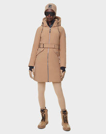 A person in a RUDSAK MILENA - 8122536 TOFFEE long down parka with a belt, tan boots, beige hat with black logo, black gloves, beige leggings, and dark sunglasses stands against a plain white background.