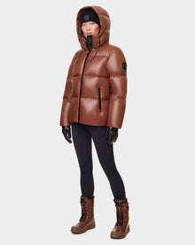 A person wears the RUDSAK DORA - 8122547 COHIBA oversized shiny brown down-filled puffer jacket with removable hood, black leggings, gloves, and brown winter boots, standing against a plain light gray background.