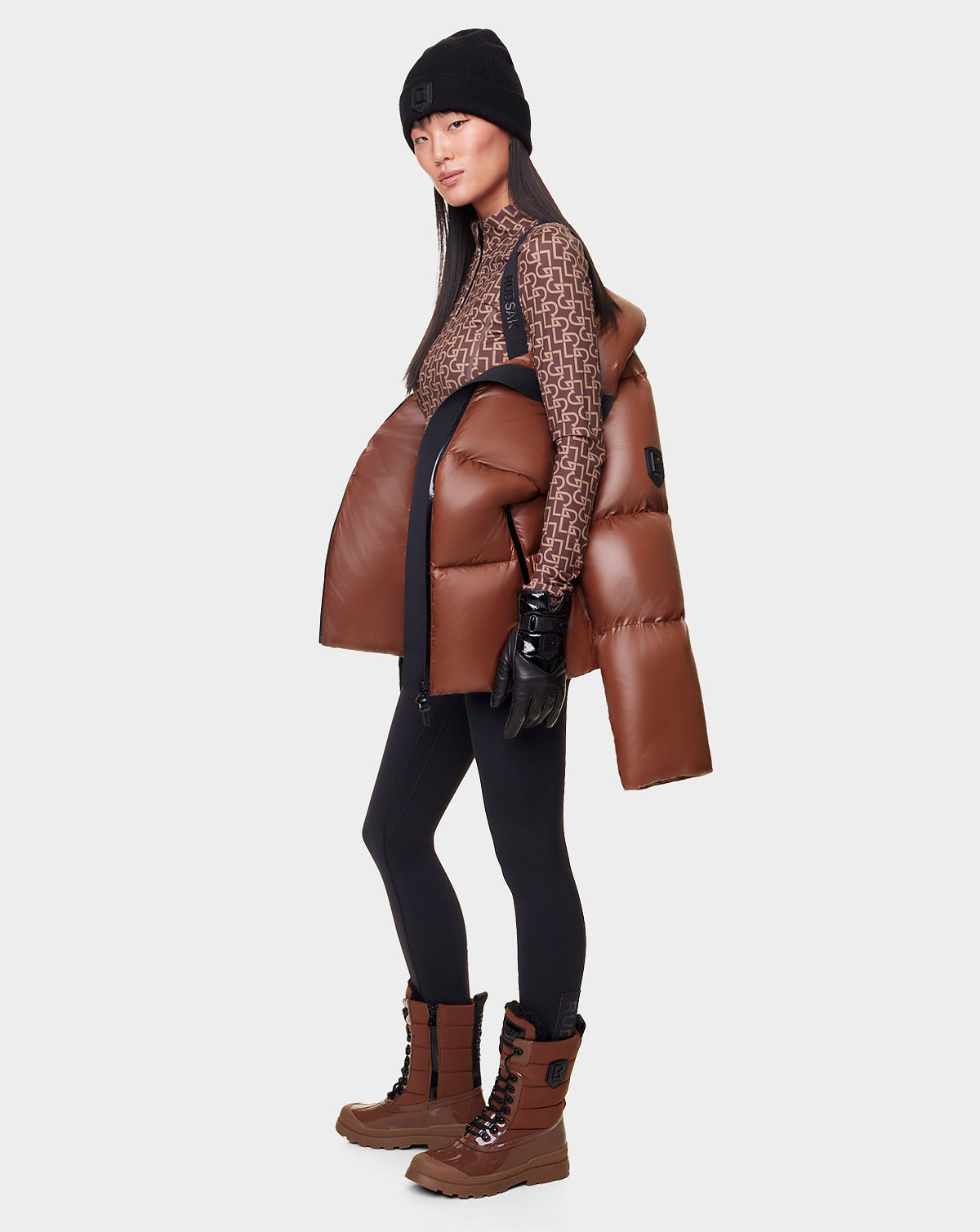 A woman wearing the RUDSAK DORA - 8122547 COHIBA down-filled women’s jacket with removable hood poses in black leggings, beanie, gloves, patterned brown turtleneck, and lace-up boots against a white background.