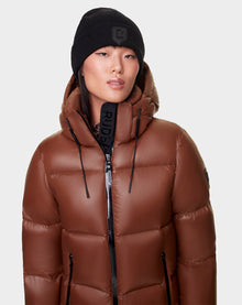 A woman in the RUDSAK DORA - 8122547 COHIBA oversized brown down-filled puffer jacket with a hood and a black beanie looks slightly to the side against a plain light background.