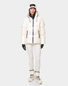 A person wearing the RUDSAK DORA - 8122547 ICELAND white down-filled women’s puffer jacket with removable hood, white leggings, winter boots, black gloves, and a white beanie stands against a light gray background.