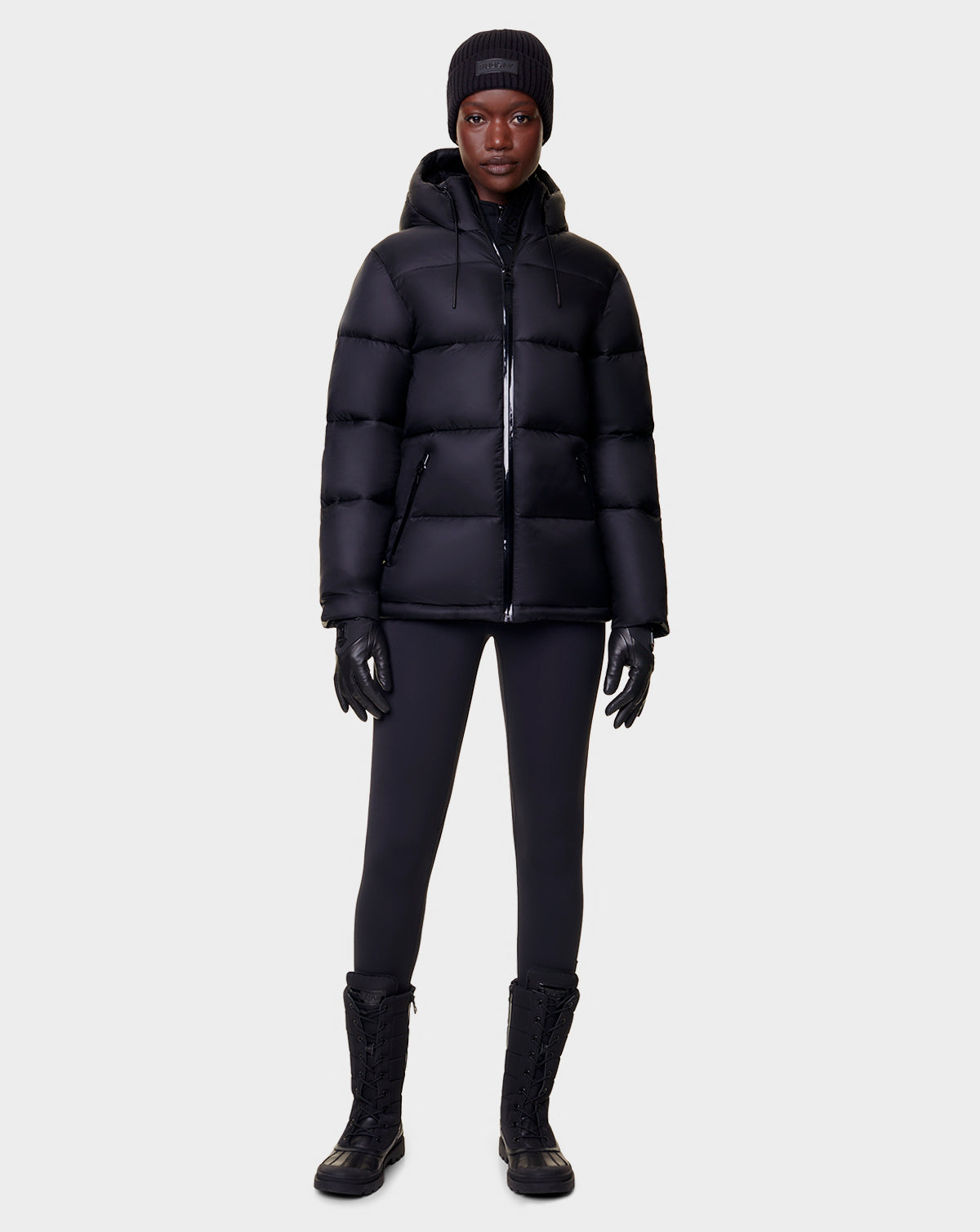 A person faces forward wearing the Rudsak EVIE - L122517 BLACK womens RDS-certified down puffer jacket, along with a black beanie, gloves, leggings, and lace-up boots against a plain light gray background.