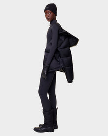 A person dressed in black winterwear stands sideways, holding the Rudsak EVIE - L122517 BLACK RDS-certified down puffer draped over one arm, against a plain light background.